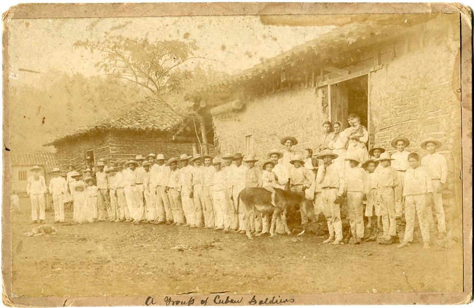 Image of group of Cuban soldiers, wearing civilian clothes, with native Cuban women, children, and elderly men posed outside of mud-brick buildings during the Cuban-Spanish revolution.