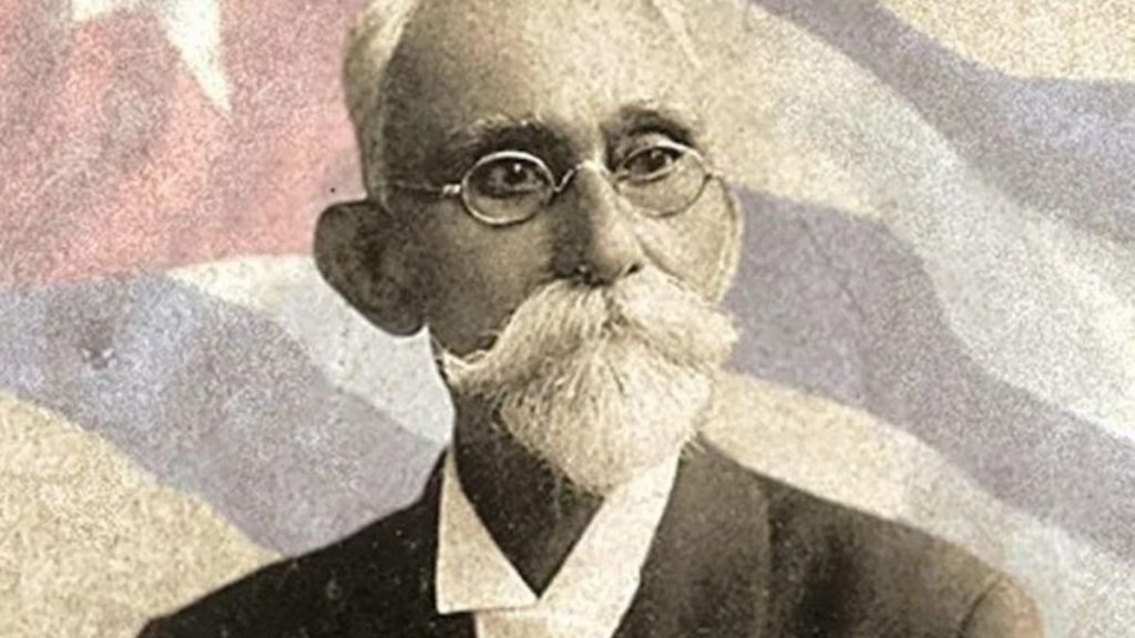 Image of an old man, Máximo Gómez, wearing glasses with a thick mustache and beard. His portrait is overlaid on an image of the Cuban flag.